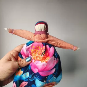 Lalka Motanka wykonana z tkanin i nici. Ma na fartuszku wielki różowy kwiat.