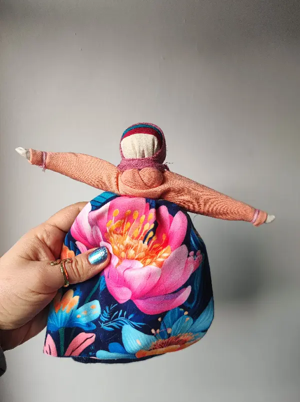 Lalka Motanka wykonana z tkanin i nici. Ma na fartuszku wielki różowy kwiat.