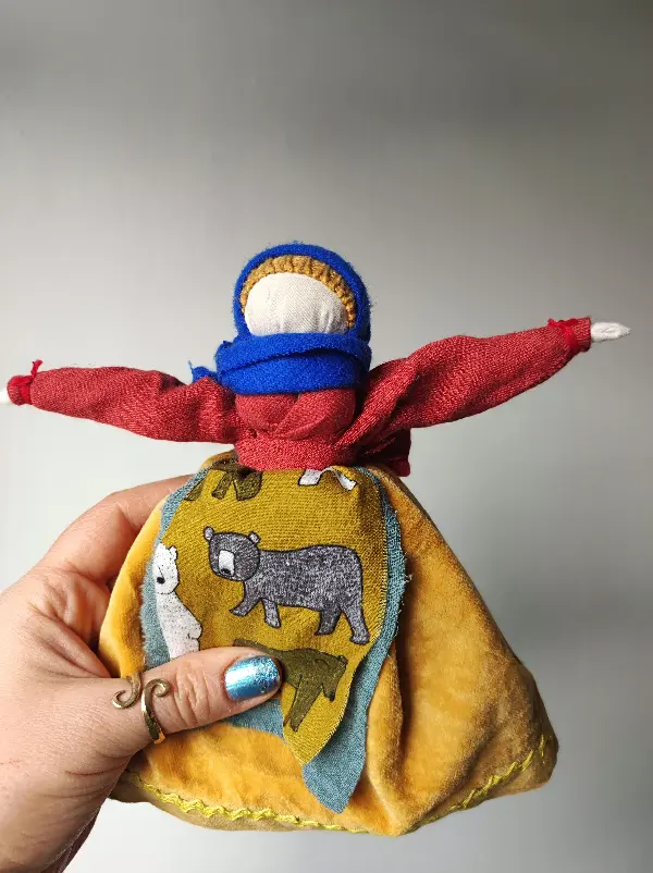 Motana Lalka Mocy wykonana z tkanin i nici. Ma na sobie fartuszek z niedźwiedziami i kobaltową chustę